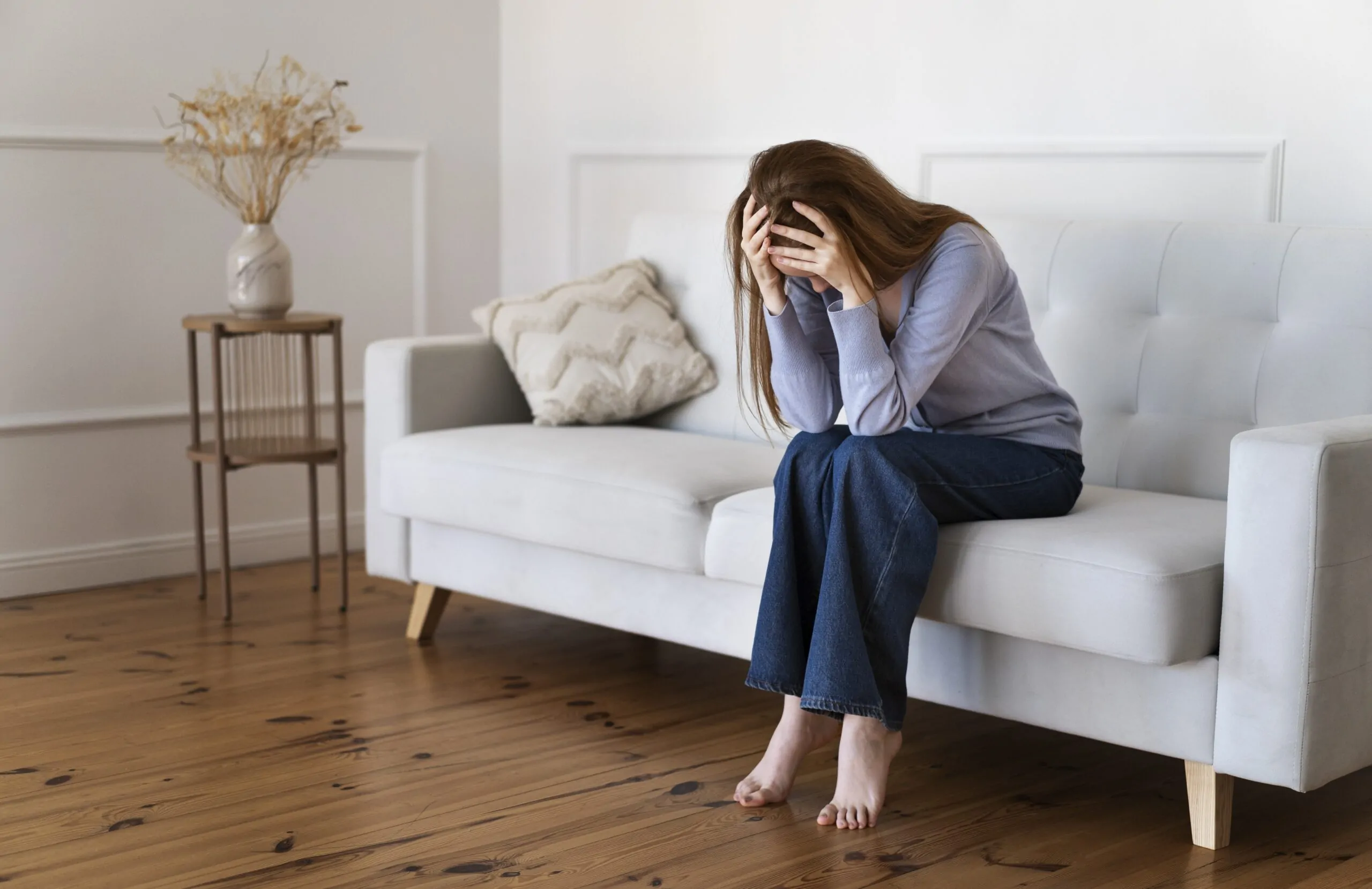 A woman sitting on a couch with her head in her hands, experiencing anxiety. Suzanne Gallagher, a Psychiatric Nurse Practitioner, offers anxiety treatment in CT with in-person appointments in Darien and New Canaan.